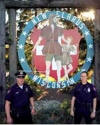 Two policemen in uniform standing before a New Glarus, Wisconsin, sign. The sign depicts Wilhelm Tell with his crossbow and son, standing before the white cross on red shield of Switzerland